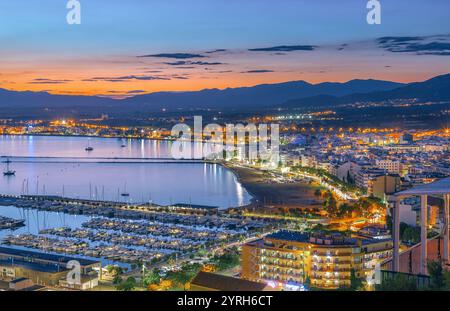 Rose, la spagna si anima al tramonto, con il caldo bagliore che illumina il paesaggio cittadino, il porto e le barche, creando una pittoresca scena di bellezza costiera Foto Stock