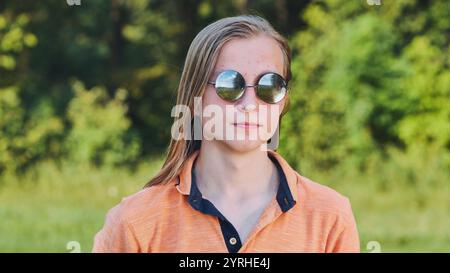 Ritratto di un adolescente biondo con i capelli lunghi che indossa gli occhiali da sole e posa in natura. Foto Stock