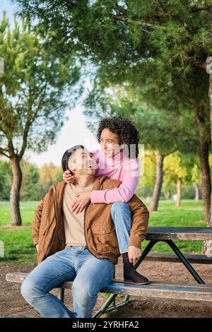 Una coppia gioiosa che ride e abbraccia su una panchina del parco circondata da lussureggianti alberi verdi, godendosi la compagnia reciproca in un ambiente naturale sereno Foto Stock