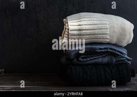 Una pila ordinata di maglioni lavorati a maglia in tonalità neutre, tra cui beige, grigio e nero, su una superficie in legno scuro, che evocano calore e intimità Foto Stock