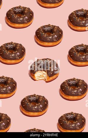 Un motivo senza cuciture di deliziose ciambelle glassate al cioccolato con spruzzette su sfondo rosa una ciambella ha un boccone allettante, creando una comicità giocosa Foto Stock