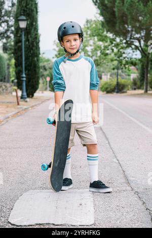 Un ragazzino si trova in una strada della città con il suo skateboard, indossando un casco per gli alberi di sicurezza lungo il percorso, creando un'atmosfera invitante Foto Stock