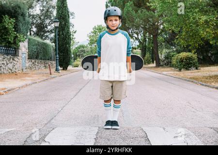 Un ragazzino si trova in una tranquilla strada di periferia, tenendo in mano uno skateboard indossa un casco e un abbigliamento sportivo casual, pronto per un avventuriero all'aperto Foto Stock