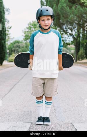 Un ragazzino si trova in una strada con uno skateboard e indossa un casco, calzini a righe e abiti casual, pronti per un'avventura in skateb Foto Stock