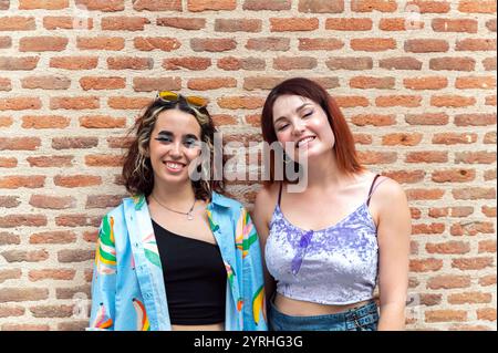 Ritratto di due giovani donne alla moda che posano felicemente contro un muro rustico di mattoni. Uno indossa una giacca colorata e occhiali da sole, l'altro un delicato p Foto Stock