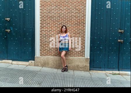 Una giovane donna con un abbigliamento casual estivo si appoggia su una parete di mattoni marroni accanto alle porte in legno blu intemprato. Foto Stock