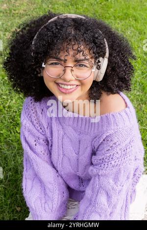 Una allegra ragazza afro-latina che indossa occhiali e cuffie, seduta sull'erba, indossa un maglione di lavanda i suoi capelli ricci e il suo sorriso brillante donano un relax Foto Stock