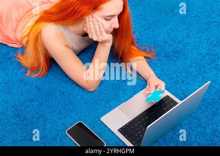 Una persona dai capelli vivaci sta sdraiandosi su un morbido tappeto blu, comodamente avvolto in una coperta che tiene in mano una carta di credito e utilizza un computer portatile Foto Stock