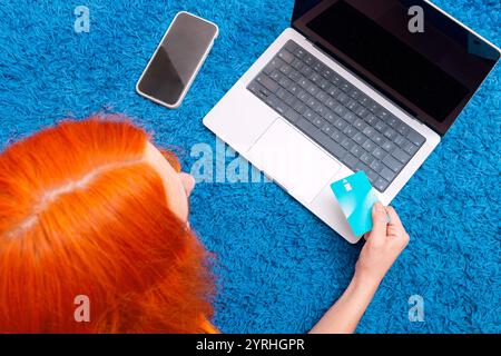 Una persona con vivaci capelli rossi fa acquisti online utilizzando un computer portatile, con una carta di credito, mentre si siede su un morbido tappeto blu Uno smartphone si trova nelle vicinanze, sugge Foto Stock