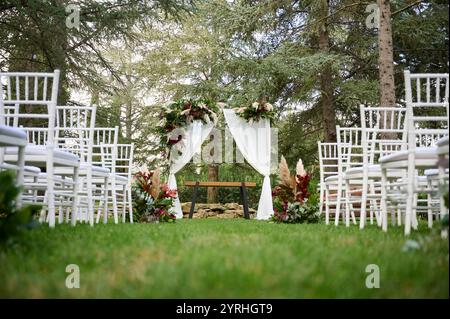 Un elegante scenario di matrimonio all'aperto caratterizzato da un arco floreale con tende bianche tra gli alberi sedie bianche fiancheggiano il corridoio erboso, creando un ambiente sereno e pictu Foto Stock