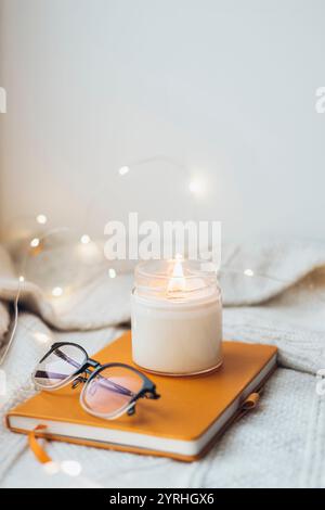 Un ambiente caldo e festoso caratterizzato da una candela illuminata, occhiali da vista e un taccuino su una morbida coperta in maglia circondata da delicate luci a corda, crea un calmo Foto Stock