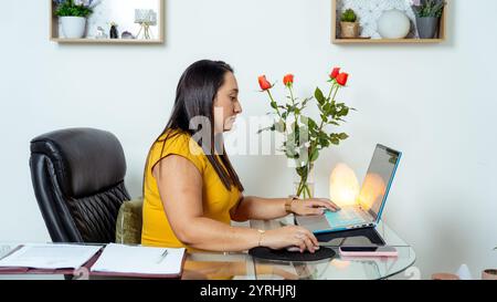 Una donna d'affari con una camicetta gialla lavora diligentemente alla sua scrivania, con i suoi scaffali per laptop con oggetti decorativi e una pianta di rose aggiungono un tocco Foto Stock