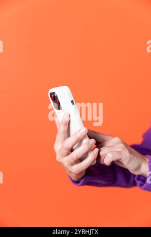 Un'immagine ravvicinata che mostra un paio di mani con uno smartphone bianco, scattata su uno sfondo arancione. Le mani sono viste in parte, enfatizzando Foto Stock