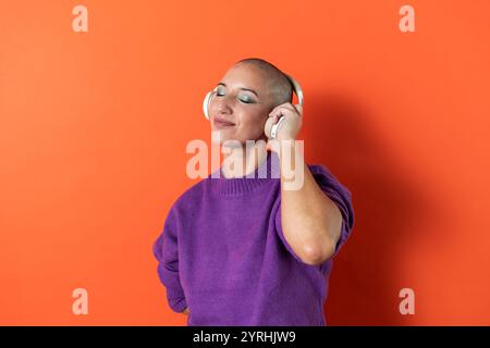 Una donna adulta serena con la testa calva, vestita con un ricco maglione viola, ascolta con gioia la musica con le cuffie bianche contro una vivace schiena arancione Foto Stock