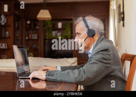 Un uomo maturo con i capelli grigi, indossa una cuffia, effettua una videochiamata su un computer portatile da casa. È concentrato e attento, mostrando una dinam di lavoro a distanza Foto Stock