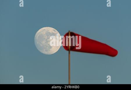 Una luna piena luminosa si innalza contro un cielo blu limpido al crepuscolo, accostata a una calza a vento rossa. La serena atmosfera serale mette in risalto la luce della luna Foto Stock