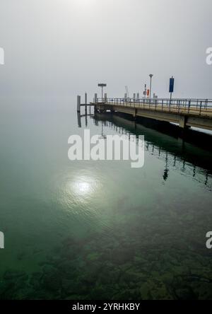 Una tranquilla scena di un molo che si estende sul Lago di Ginevra in una giornata nebbiosa, le acque calme rispecchiano il cielo tenue e creano un'atmosfera serena e tranquilla Foto Stock