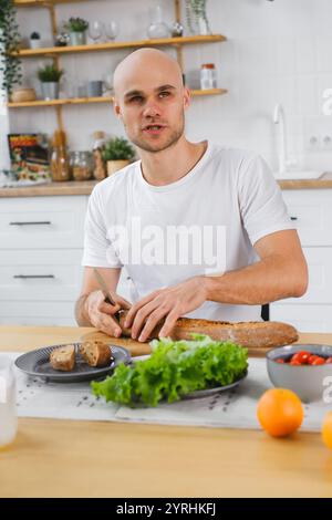 Un cieco affetta il pane a un tavolo da cucina, circondato da ingredienti freschi, tra cui lattuga e pomodori. L'ambiente include una moderna e ben-orga Foto Stock