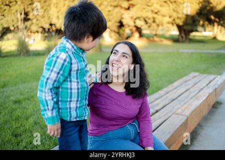 Un momento gioioso tra madre e figlio nel parco la madre, seduta su una panchina di legno, guarda con amore il suo bambino, che è in piedi accanto a lei dentro Foto Stock