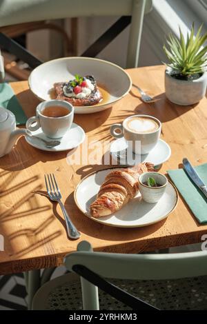 Tavolo da colazione illuminato dal sole con croissant, latte cremoso, tè caldo e una ciotola di frutta fresca, Un ambiente incantevole con luce naturale e vegetazione Foto Stock