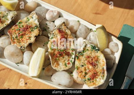 Un primo piano di appetitose ostriche al forno guarnite con erbe e croccanti pangrattati, servite con spicchi di limone su un piatto decorativo in pietra Perfect gou Foto Stock