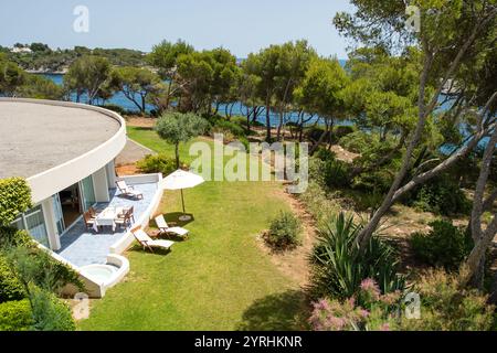 Scoprite una moderna camera d'hotel con una spaziosa area esterna con sedie a sdraio, un tavolo da pranzo e ombrelloni circondati dal verde lussureggiante e dal mare Foto Stock