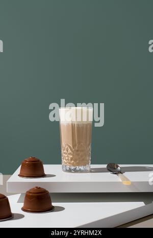 Un alto bicchiere di caffè ricoperto di crema di latte, su un tavolo moderno con capsule di caffè e un cucchiaio nelle vicinanze, perfetto per immagini a tema caffetteria Foto Stock