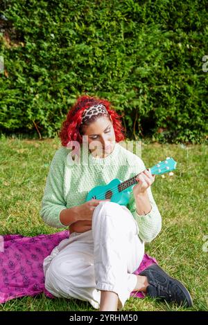Donna trans con i capelli rossi suona un ukulele di tè in un parco tranquillo con un maglione verde e pantaloni bianchi, siede sull'erba, godendo di una moma tranquilla Foto Stock