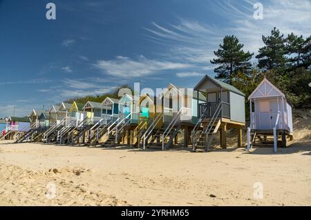 Una fila di colorate capanne da spiaggia su una spiaggia sabbiosa, sotto un cielo blu con nuvole sparse, le affascinanti cabine in legno forniscono un vibrante contrasto con il loro Foto Stock