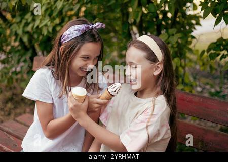 Due ragazze allegre sedute su una panchina in un parco, ridendo e condividendo i coni del gelato, la vegetazione lussureggiante e le espressioni gioiose catturano un divertimento e una spensieratezza Foto Stock