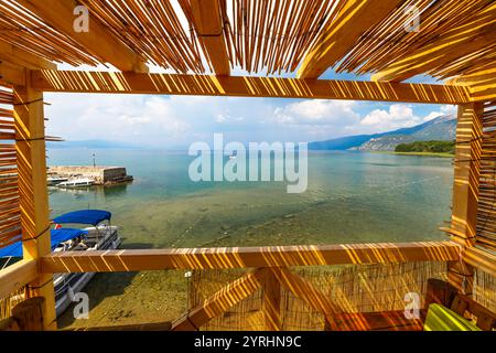 La luce del sole filtra tra le spranghe di legno di una capanna sulla spiaggia, rivelando un panorama mozzafiato sulle acque cristalline del lago di Ocrida, sulle barche e sulle montagne lontane Foto Stock