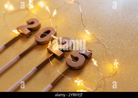 Le candele del Capodanno d'oro 2025 con le luci delle stelle delle ghirlande si trovano su uno sfondo dorato. Carta da parati per le feste Foto Stock