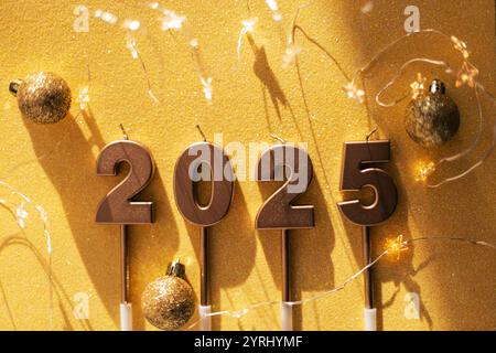 Le candele del capodanno 2025 dorate con stelle, luci e palline dorate si trovano su uno sfondo dorato. Carta da parati magica per le feste Foto Stock