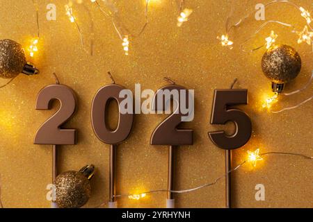 Le candele dorate festive 2025 con stelle luminose giacciono con palline dorate su sfondo dorato. Carta da parati per le vacanze. Capodanno 2025 e cartolina di Natale Foto Stock