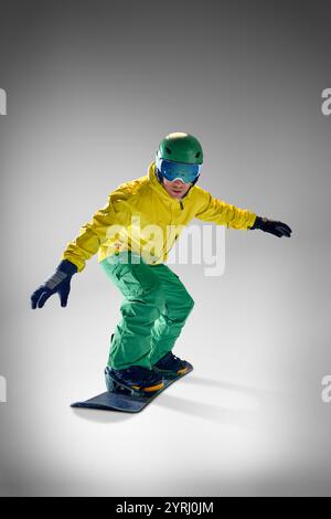 Snowboarder, giovane atletico, con giacca gialla e pantaloni verdi che eseguono trick a mezz'aria su sfondo grigio studio. Foto Stock