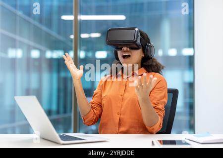 Giovane donna d'affari che utilizza un visore per la realtà virtuale in ufficio. È seduta a una scrivania con un portatile, esprimendo entusiasmo e stupore. Tecnologia e innovazione sono concetti evidenziati. Foto Stock