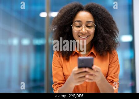 Giovane donna d'affari che sorride usando il suo tphone. Sembra coinvolta e soddisfatta del suo dispositivo, catturando un momento di produttività e connessione in un ambiente di ufficio moderno. Foto Stock