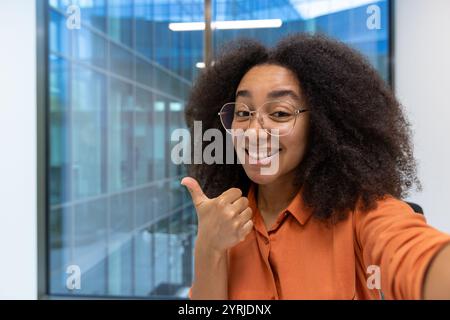 Giovane donna d'affari con una camicia arancione che sorride e si fa un pollice. Si trova in un ufficio luminoso e moderno, che trasmette positività e fiducia. Foto Stock