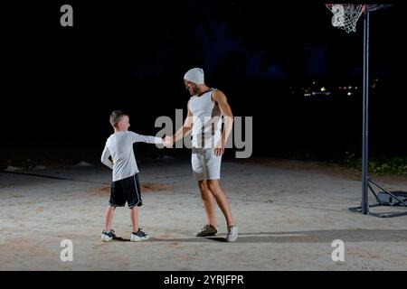 Un padre gioca a basket con il suo giovane figlio di notte in un parco illuminato da una luce brillante Foto Stock