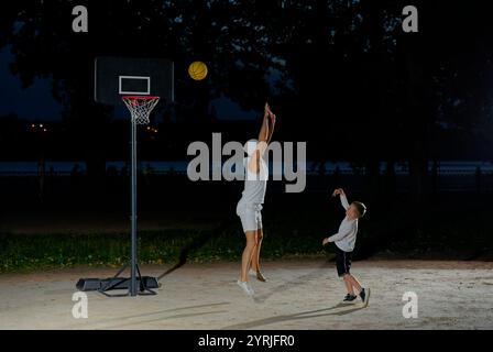 Un padre gioca a basket con il suo giovane figlio di notte in un parco illuminato da una luce brillante Foto Stock