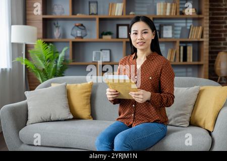 Donna asiatica in outfit casual, seduta sul divano tenendo documenti, sorridendo. Ambiente domestico con atmosfera accogliente, libreria e piante. Il comodo spazio abitativo riflette il relax e la calma. Foto Stock