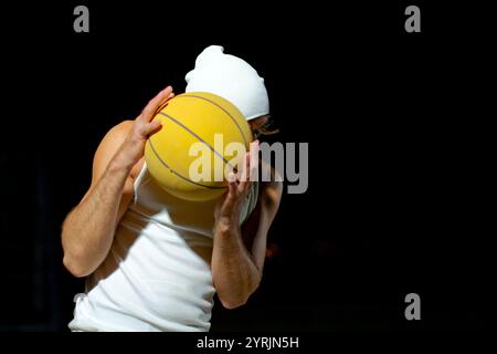 Un uomo vestito con canotta bianca e berretto crea una posa dinamica con una pallacanestro gialla. La sua espressione sicura mette in risalto la sua passione per il Foto Stock