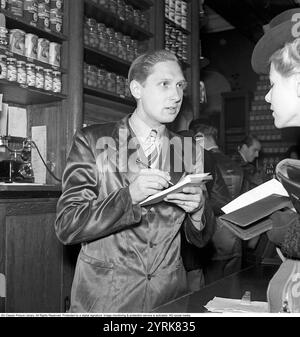 Nel negozio nel 1942. Un assistente maschile nella gastronomia di Arvid Nordquist a Birgerjarlsgatan a Stoccolma. Siamo nel 1942 e la seconda guerra mondiale sta infuriando in Europa. Nonostante la guerra, l'azienda è in grado di offrire generi alimentari e prelibatezze. Il negozio non è ancora completamente passato al self-service, ma qui la giovane cliente donna si trova con la sua lista della spesa. L'assistente maschile ascolta attentamente e prende appunti. Mentre il cliente attende, preleva i prodotti desiderati e li imballa. 1942. Kristoffersson rif. A126-6 Foto Stock