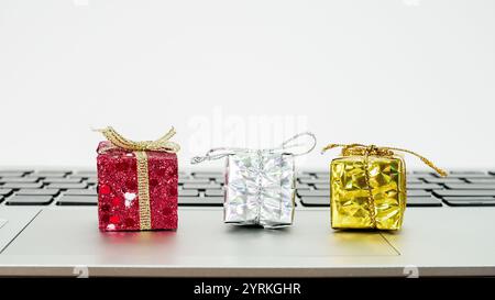 Tre minuscole confezioni regalo in rosso, argento e oro si trovano in cima alla tastiera di un notebook, aggiungendo un tocco di festa a un ambiente natalizio. Foto Stock