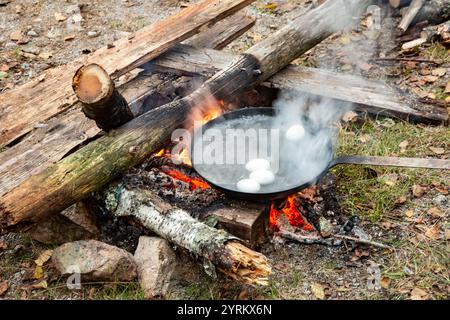 Primo piano del fuoco rosso a legna acceso all'aperto con una padella riempita di acqua bollente in cui le uova vengono cotte come parte dell'autosufficienza Foto Stock
