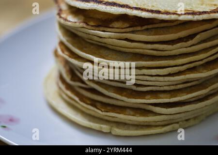 Un'alta pila di sottili pancake marrone chiaro si trova su un piatto bianco con un sottile design floreale. I pancake sono impilati in modo ordinato, mostrando un po' marroni Foto Stock