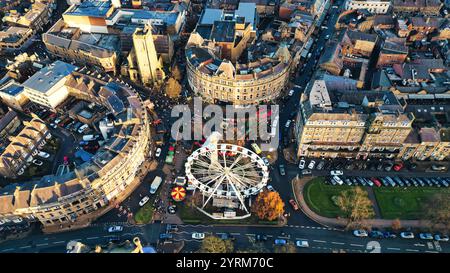 Vista aerea di una piazza della città con una ruota panoramica, circondata da edifici, auto parcheggiate e pedoni. Gli edifici sono principalmente a più piani e ston Foto Stock