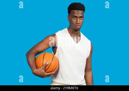 Ritratto di un giovane giocatore di basket sicuro di sé che regge un pallacanestro su uno sfondo blu vibrante Foto Stock