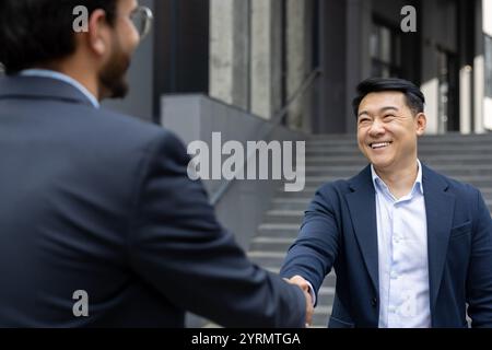 Due professionisti in abbigliamento formale stringono la mano, simboleggiando una partnership di successo. L'ambiente esterno e i sorrisi luminosi trasmettono fiducia, connessione e collaborazione. Foto Stock