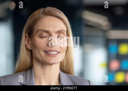 Donna matura donna d'affari sembra calma e concentrata, occhi chiusi, in un ambiente moderno di ufficio. La sua espressione serena suggerisce consapevolezza e relax, ideale per illustrare l'equilibrio e l'armonia sul posto di lavoro. Foto Stock